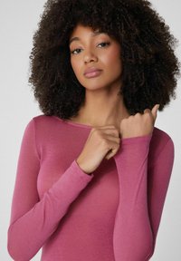 Přiléhavý top s dlouhým rukávem z jemné růžové tkaniny, s kulatým výstřihem a hladkým povrchem. Modelka je pózována s rukama blízko obličeje.