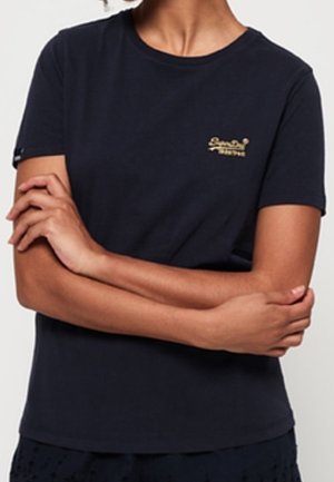 Osoba ubrana w dopasowaną czarną koszulę z krótkim rękawem z małym złotym logo na piersi, z założonymi rękami na wysokości talii.