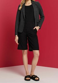 Kvinna iförd svarta shorts, svart topp, mörkgrå blazer, svarta sandaler och gyllene armband som står mot en röd och vit bakgrund.