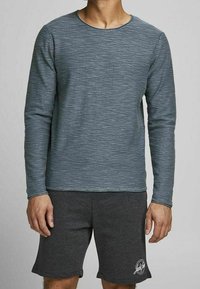 Homme portant une chemise grise à manches longues texturée et un short gris foncé avec un logo rond sur la jambe gauche, debout devant un fond uni.