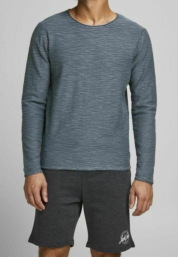 Homme portant une chemise grise à manches longues texturée et un short gris foncé avec un logo rond sur la jambe gauche, debout devant un fond uni.