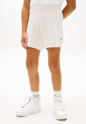 Vrouw draagt witte gebreide shorts, witte sokken met merklogo en witte hoge sneakers, staande tegen een effen achtergrond.