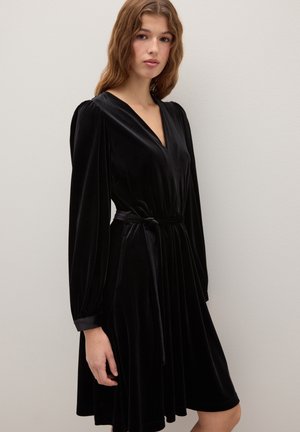 Vestido cruzado de terciopelo negro con mangas largas, escote en V y cintura con cinturón. Presenta detalles sutiles recogidos en los hombros y falda fluida.