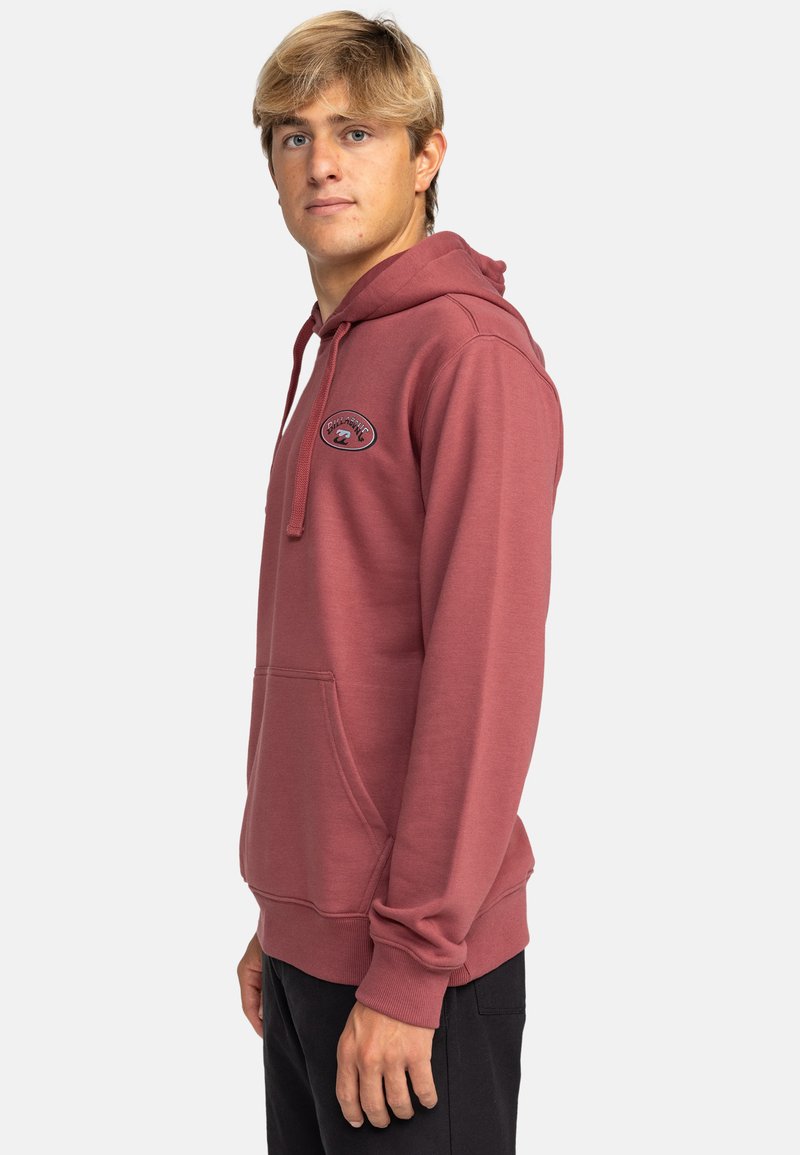 Joven con pelo rubio que lleva una sudadera burdeos con bolsillo delantero y pantalones negros, de pie contra un fondo claro y liso.