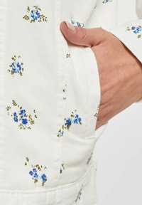 Hvitt stoff med et blomstermønster av små blå blomster og grønne blader. Hånd hvilende i en sidelomme, som viser teksturen av materialet.