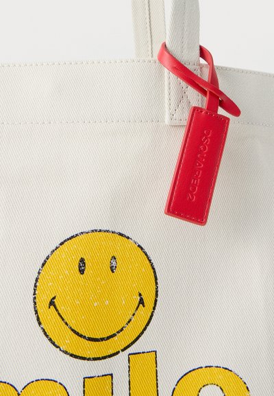 Μπεζ υφασμάτινη τσάντα με μεγάλο φθαρμένο κίτρινο χαμογελαστό πρόσωπο και κόκκινη δερμάτινη ετικέτα με ανάγλυφη την επιγραφή "DSQUARED2" στο χερούλι.