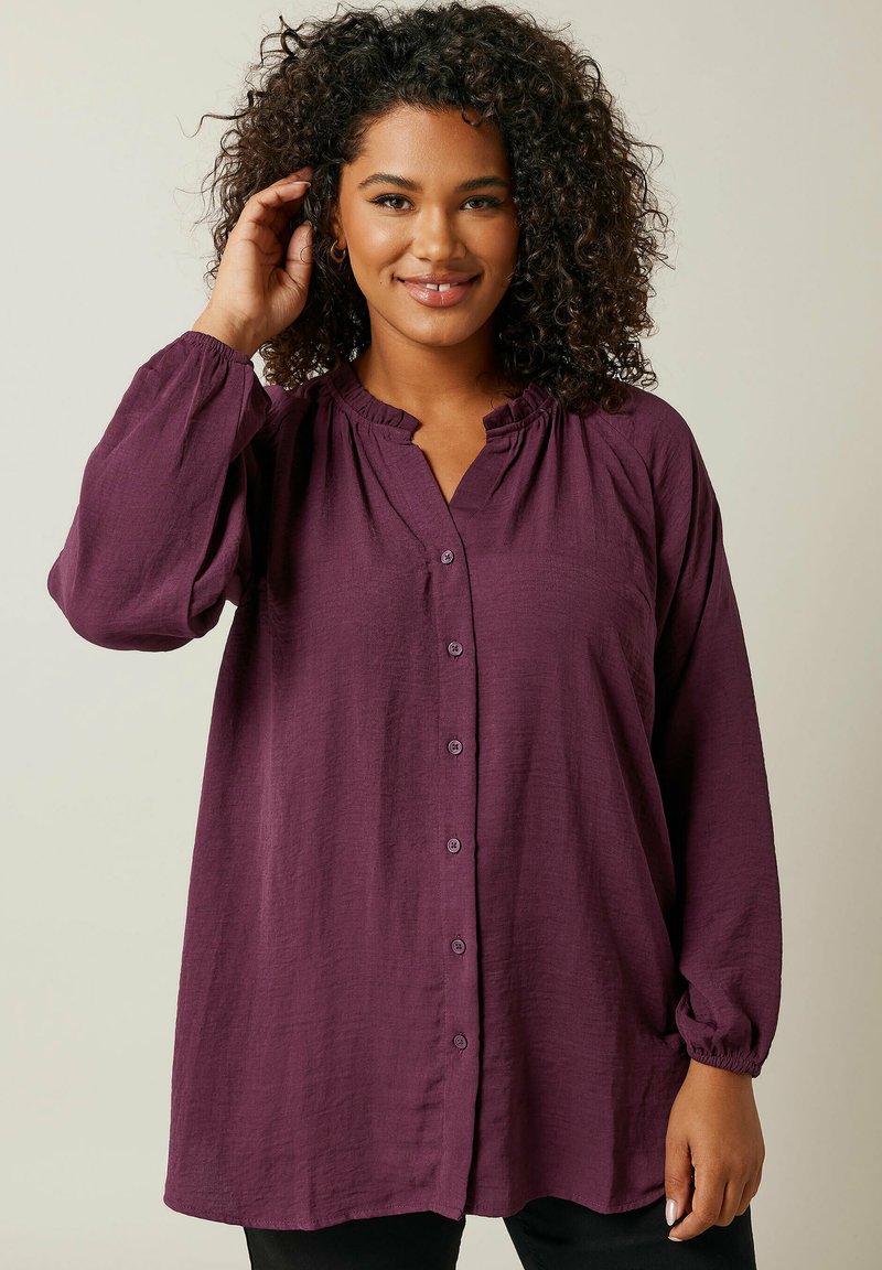 Blusa viola a maniche lunghe con vestibilità comoda, chiusura con bottoni sul davanti, scollatura arricciata e tessuto texturizzato. Presenta polsini elasticizzati e una morbida caduta.