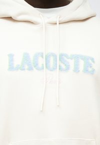 Sweatshirt com capuz creme com um logótipo "LACOSTE" felpudo multicolorido em azul e verde, com "Paris" em rosa claro por baixo. Cordões ajustáveis.