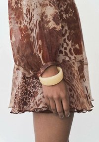 Fließendes, transparentes Kleid mit braunem und cremefarbenem Tiermuster, das puffige Ärmel und einen gerafften Saum hat. Am Arm befindet sich ein glattes, blassgelbes Armband.