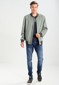 Svetlošedá bomber bunda s rebrovanými manžetami a golierom, kombinovaná so zničenými modrými džínsy a šedými teniskami. Na bunde nie sú viditeľné logá.