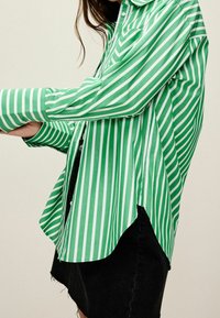 Kobieta w zielono-białą pionowo paskowaną koszulę z guzikami i czarną dżinsową spódnicą, stojąca na jednolitym tle.