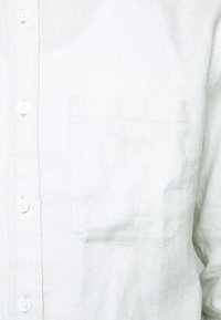 Ljusgrön linnekavaj med bröstficka, vita knappar och en strukturerad tygkvalitet. Enkel, ren design.