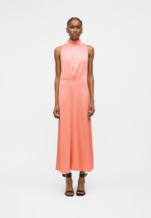 Kobieta stojąca w bezrękawnym, z wysokim kołnierzem, sięgającej kostek koralowo-różowej sukience oraz czarnych, otwartych sandałkach na wysokim obcasie na białym tle.