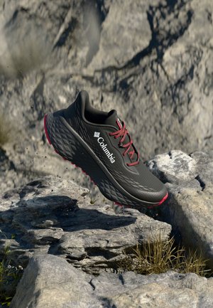 Schwarzer Columbia Trailrunning-Schuh mit roten Schnürsenkeln und Sohle, schwebt über felsigem Gelände im Freien.