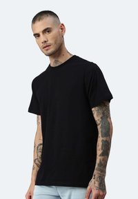 Schwarzes Baumwoll-T-Shirt mit kurzen Ärmeln und Rundhalsausschnitt, getragen von einer tätowierten Person. Weiche Textur und normale Passform.