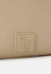Sac beige avec une surface texturée et un logo "recycler" en relief sur un patch rectangulaire, avec des coutures ton sur ton délimitant le patch.
