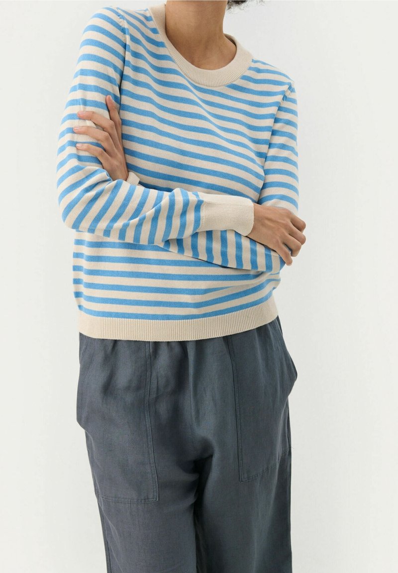 Personne portant un pull rayé bleu clair et beige à manches longues et un pantalon gris foncé, bras croisés devant.