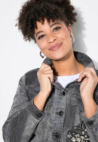 Grå denimjacka med en texturerad design, knäppning med knappar och blommönsterbroderi på vänster sida. Med krage och en varierad mönsterfinish.