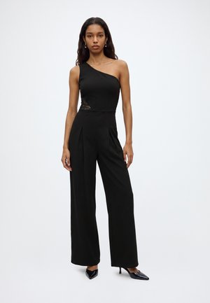 Vrouw draagt een zwarte jumpsuit met één schouder, wijde pijpen en zwarte hakken, staand tegen een effen witte achtergrond.