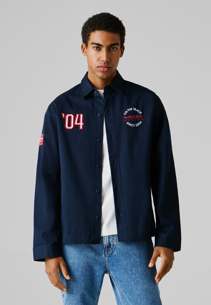 Νεαρός άνδρας που φοράει μπλε σκούρο πουκάμισο με κουμπιά με τα σύμβολα "’04" και το λογότυπο της Red Bull Racing, λευκό πουκάμισο από μέσα και μπλε τζιν, σε απλό φόντο.