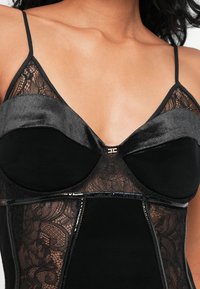 Μαύρα εσώρουχα με δαντελένιες λεπτομέρειες, διαθέτοντας επενδεδυμένα cups, λεπτές τιράντες και κεντρική χρυσή λεπτομέρεια. Συνδυάζονται υφή δαντέλας και λεία αδιαφανή ύφασμα.