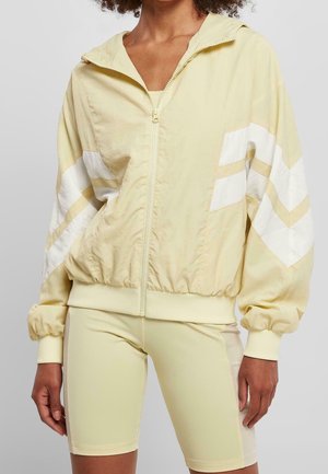 Personne portant une veste zippée jaune pâle avec des bandes chevron blanches sur les manches et un short de cycliste jaune pâle assorti.