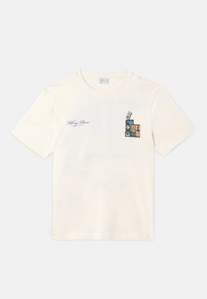 T-shirt en coton couleur crème présentant un graphique de cassettes empilées sur la poitrine gauche, avec des accents bleus et marron et du texte.
