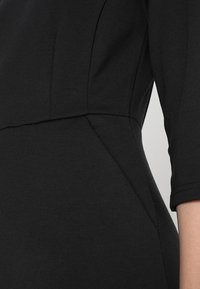 Close-up van het middenstuk van een zwart aansluitend jurkje en de rechterarm met driekwartmouwen, waarbij de naaddetails en de gladde stofstructuur zichtbaar zijn.