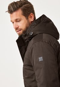 Dunkelbraune Kapuzenjacke mit strukturiertem Finish, ausgestattet mit einem Logopatch am Ärmel, Druckknöpfen und Steppdetails an der Kapuze.