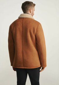 Brązowa kurtka z faux shearling z kremowym kołnierzem, o kwadratowym kroju, gładkiej fakturze i widocznych detalach szwów na plecach.
