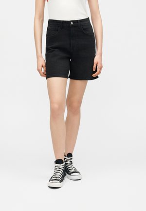 Person iført sorte denimshorts, hvid ærmeløs top og sorte højtop-sneakers, stående mod en ensfarvet hvid baggrund.