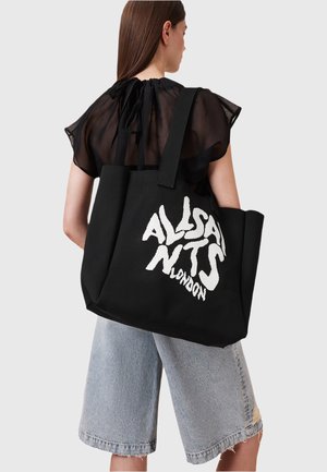 Donna con lunghi capelli castani indossa un top nero trasparente e pantaloni shorts di jeans fino al ginocchio, mentre porta una grande borsa nera con testo bianco in grassetto.