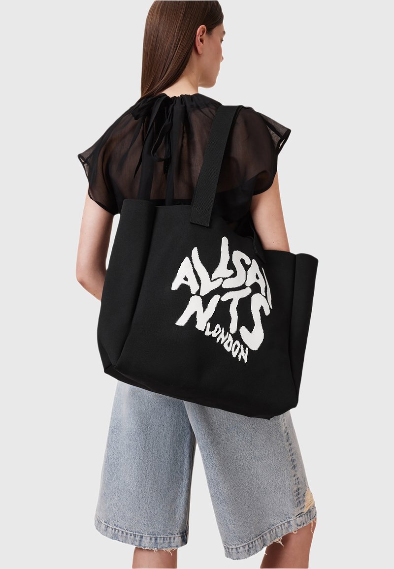 Femme aux longs cheveux bruns portant un haut noir transparent et un short en denim à mi-cuisse, portant un grand sac fourre-tout noir avec un texte blanc audacieux.