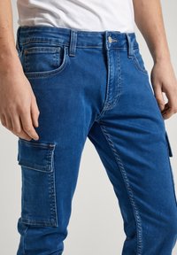 Pantalones cargo de mezclilla azul que presentan un bolsillo frontal, un bolsillo lateral y costuras en contraste. La tela tiene un acabado suave, ligeramente texturizado.