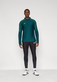 Μακριά αθλητική μπλούζα σε τιρκουάζ με φερμουάρ στο 1/4 και πράσινες λεπτομέρειες, συνδυασμένη με μαύρο παντελόνι με στενή γραμμή και υφασμένα τελειώματα, καθώς και λευκά αθλητικά παπούτσια.