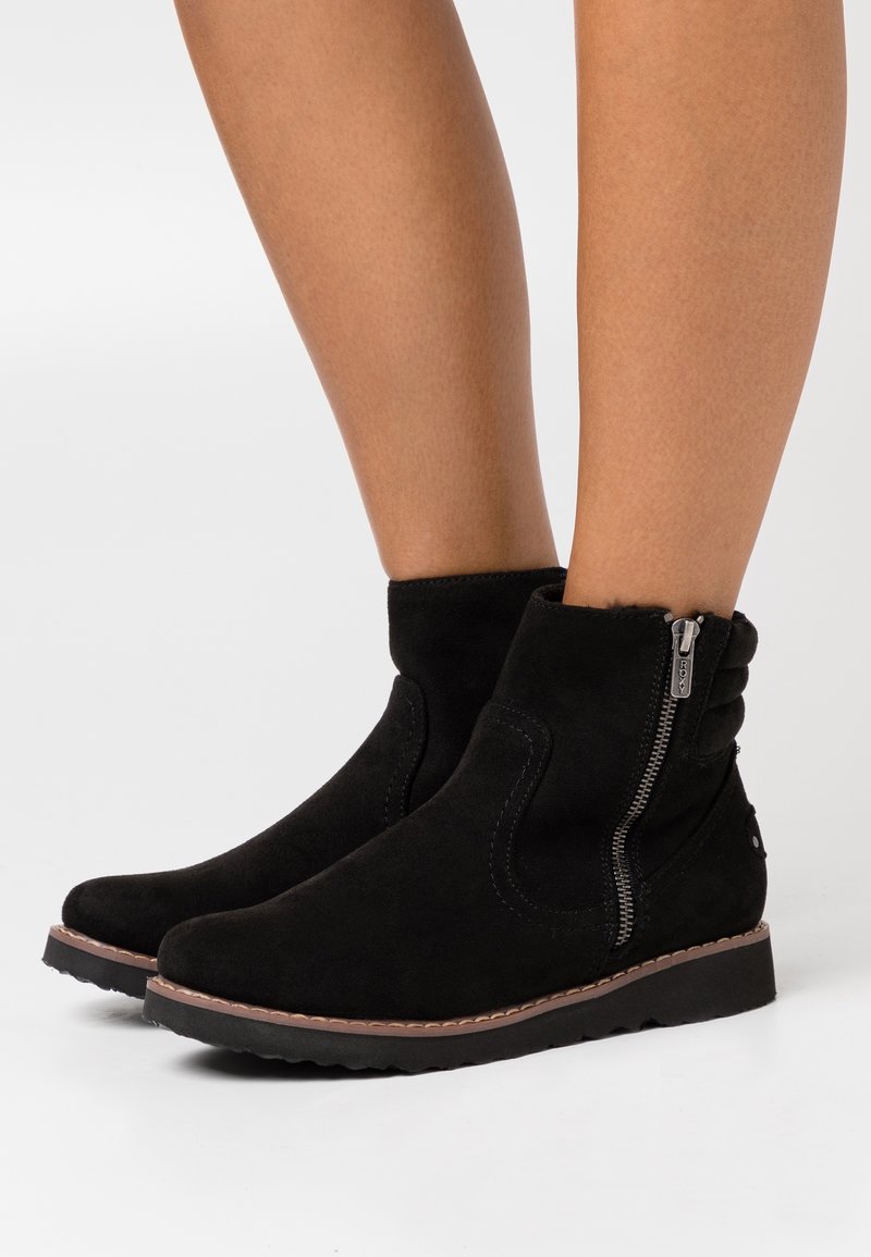 Bottes en suède noir montant à la cheville avec fermetures éclair latérales, portées sur des jambes nues, avec une semelle marron cousue et un talon léger.