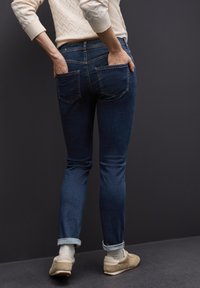 Persona vestida con jeans oscuros doblados y zapatillas beige, de pie con las manos en los bolsillos traseros contra un fondo gris oscuro.