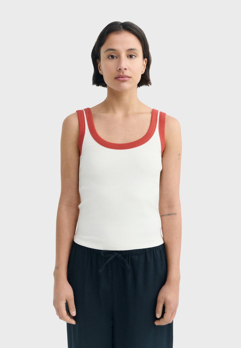 Jeune femme aux cheveux courts et foncés portant un haut blanc sans manches avec un liseré rouge et un pantalon noir, debout devant un fond uni.