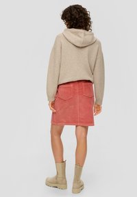 Femme se tenant de dos, portant un sweat à capuche beige, une jupe en velours côtelé rose avec des poches arrière, et des bottes chevilles beiges sur fond gris.