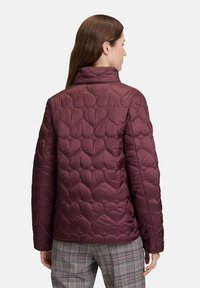 Donna che indossa una giacca trapuntata bordeaux con motivi a cuore, abbinata a pantaloni a quadri grigi, vista da dietro.
