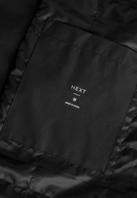 Intérieur de veste en satin noir avec une étiquette carrée en tissu indiquant "NEXT", taille "M" et la mention "MADE IN CHINA" en blanc.