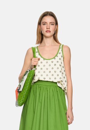 Woman wearing a sleeveless white top with green trim and floral print, green skirt, holding a green woven bag with patterned scarf.