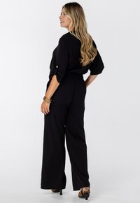 Vrouw met lang blond haar in een zwarte top met opgerolde mouwen en wijde broek, met een leopardenprint hakken en gouden armbanden, staat zijwaarts.