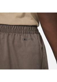 Elastický pás na hnědých kalhotách s logem "DRI-FIT", nošeno s béžovým topem a viditelnou tmavou rukou.