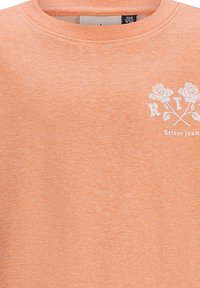 Felpa color arancio chiaro realizzata in tessuto morbido. Presenta una grafica bianca di rose e le lettere "R T", con un colletto a costine. Design semplice.