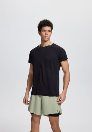 Młody mężczyzna stoi, ubrany w czarną sportową koszulkę z krótkim rękawem oraz jasnooliwkowe krótkie spodenki sportowe na jednolitym tle.