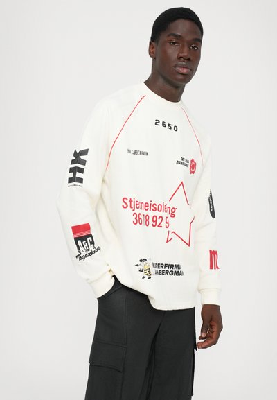 Man wearing a white long-sleeve shirt with red and black text, graphics, and numbers, paired with black cargo pants, standing against white background.