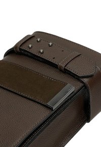 Brązowa skórzana torba z gładką fakturą, ciemnym materiałowym akcentem, metalowymi ćwiekami i srebrną płytką z logo. Posiada bezpieczne zapięcie na pasek.