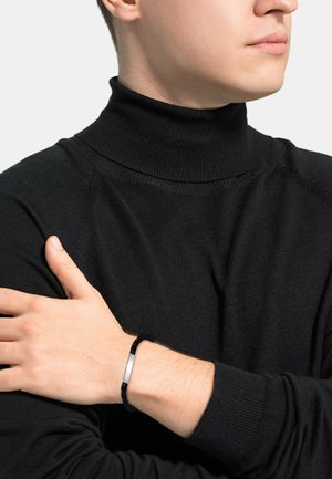 Czarny prążkowany sweter z golfem o dopasowanym kroju; modelka ma na sobie czarną bransoletkę o fakturze z srebrnym akcentem.