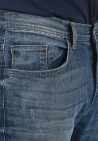 Tamno plavi traperice s leđnim džepom s detaljima šivanja, izblijelim teksturom i konvencionalnim zatvaranjem na dugme i patentni zatvarač.